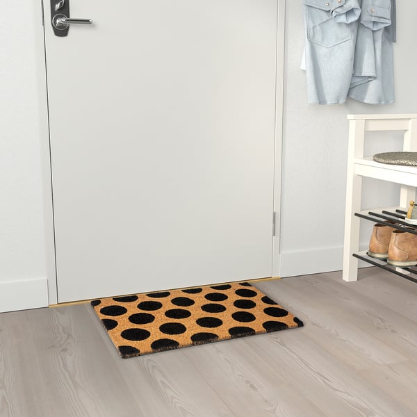 A rectangular doormat with black dots sits by a door. Its coir material and pattern make it modern and functional. Its backing keeps it in place.