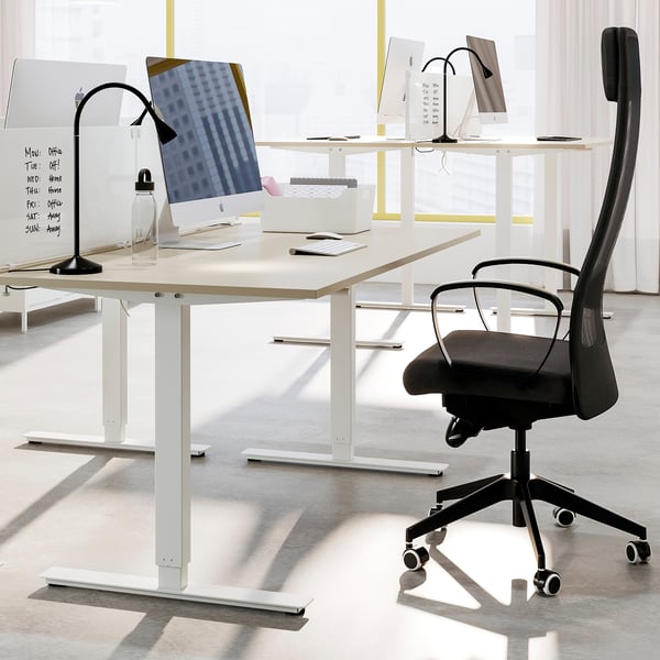 Modern office setup with white standing desks, black office chairs, and computer monitors. Desks have white crank handles and adjustable feet.