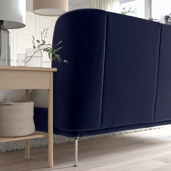 A modern bed with a dark blue, curved headboard and wooden legs. A small table beside it holds a lamp and a plant.