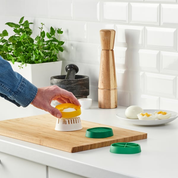 Person uses colourful egg slicer on wooden board. Modern kitchen with plant.