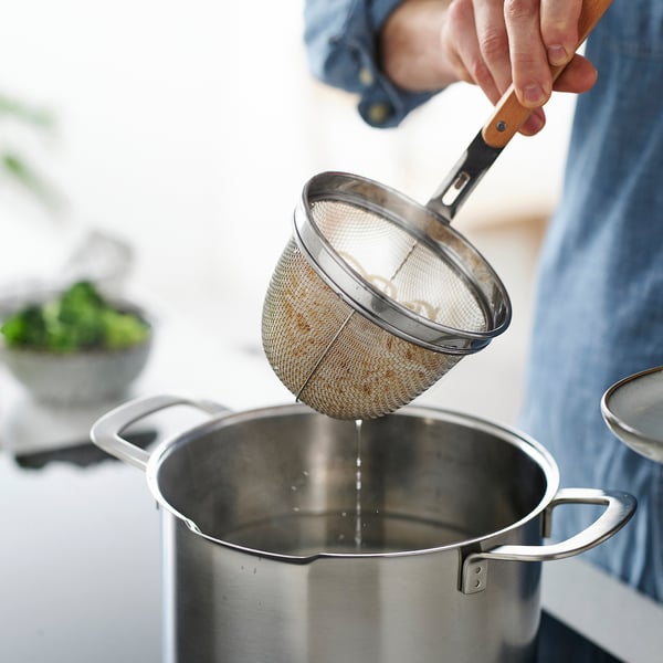 A person is draining cooked food using a VARDAGEN metal strainer with a wooden handle, showcasing its deep design and functionality.
