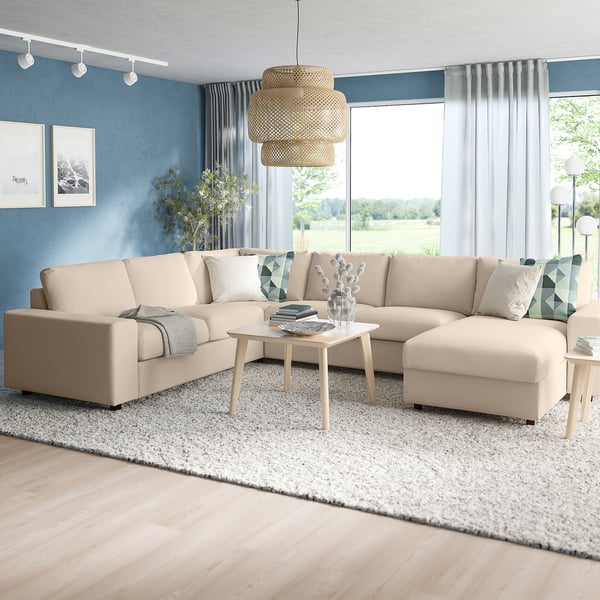 Minimalist living room: corner beige sofa, beige rug, light blue walls, natural pendant lamp. Large windows offer a garden view.