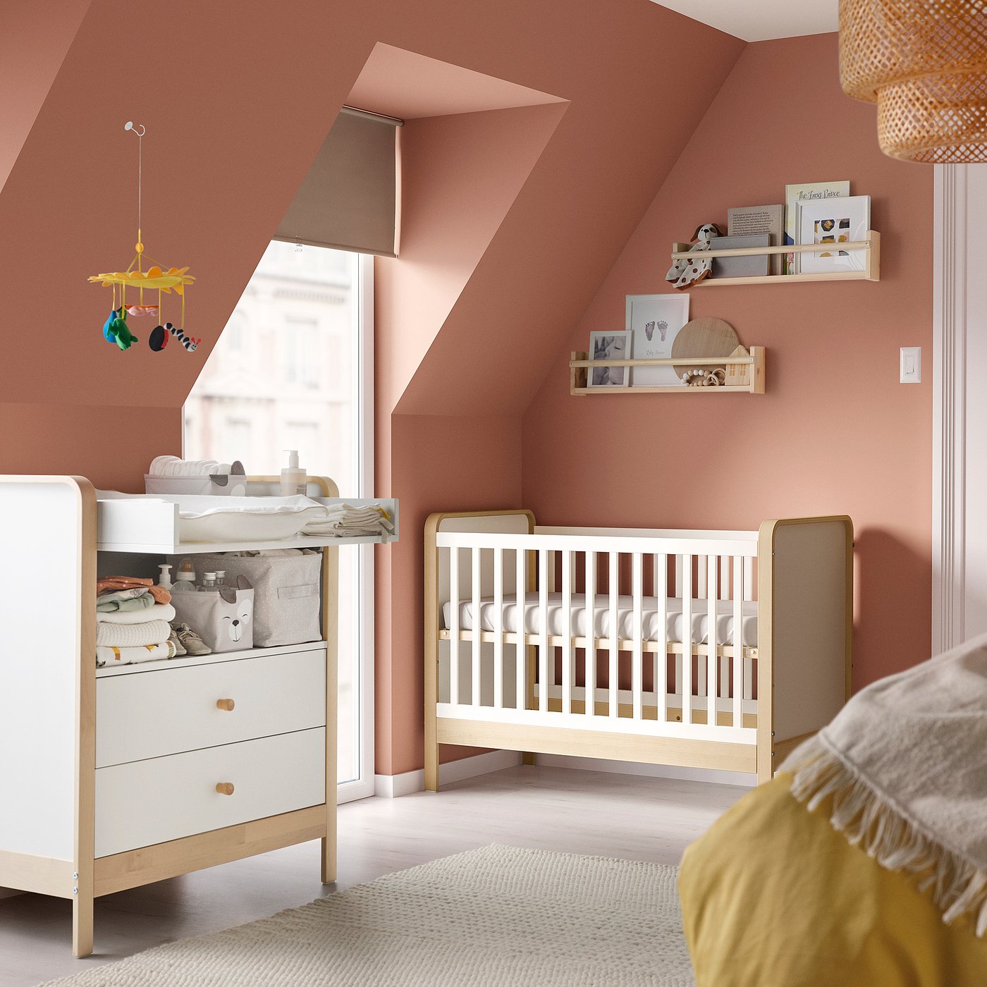 Baby nursery with pink walls, white crib, changing table, yellow mobile, shelves with toys, and beige carpet.