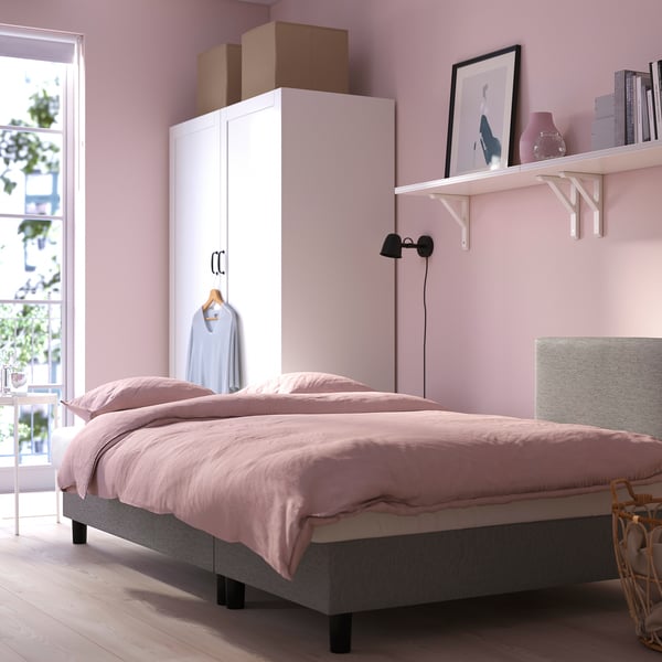 A cosy bedroom with a light pink bedspread and pillows, white shelves holding books and a vase, and a large wardrobe.