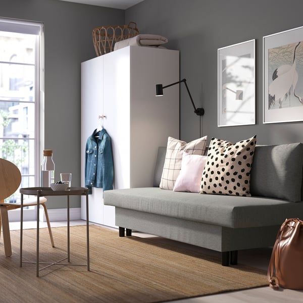Modern living room with grey sofa bed, denim jacket, white cabinet, side table, and wall art.