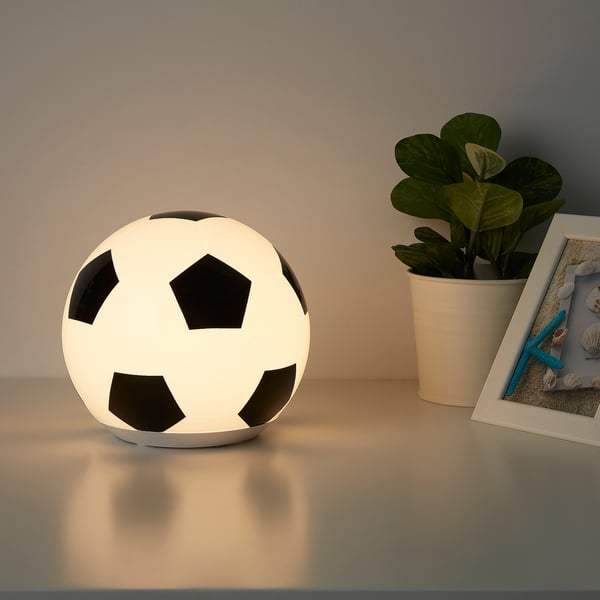 A spherical football-shaped lamp sits on a table, emitting a soft glow. Beside it are a potted plant and a framed photo.