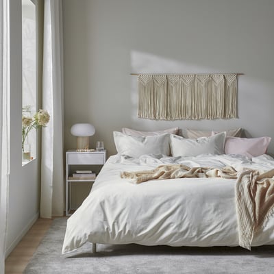 Cosy bedroom with beige bedding, pillows, bedside table, lamp, flowers, and macrame wall hanging.