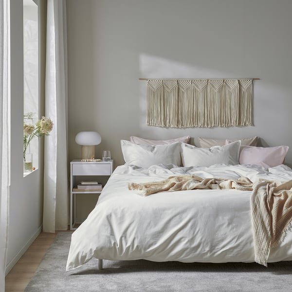 Minimalist bedroom with ÄNGSLILJA duvet, throw, bedside lamp, flowers, macrame, and natural light.
