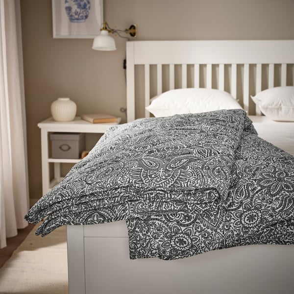 Grey & white floral duvet, ÄNGSSTAMFLY, on large bed. Bedside table and lamp beside.