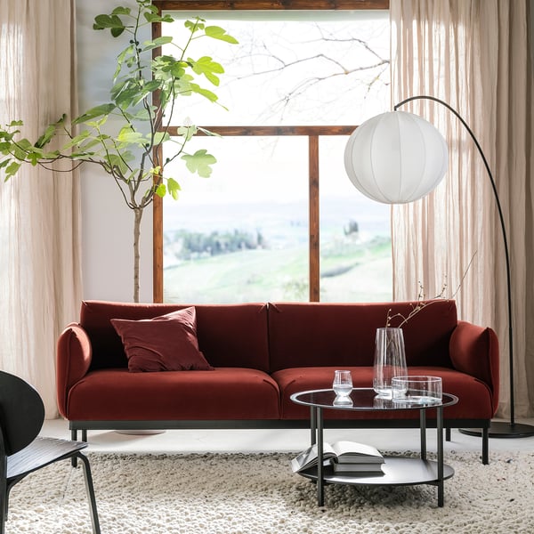 Modern living room with rust red ÄPPLARYD sofa, glass coffee table, floor lamp, and large window with view.