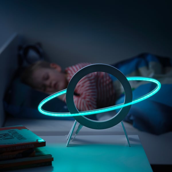 A child sleeping nearby, modern space-themed led lamp in cyan light on bedside, books stacked.