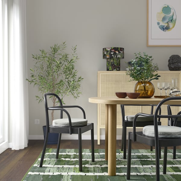 Modern dining area with black chairs, light wood table, green patterned rug, and decorative plants. Painting on wall, curtains on window.
