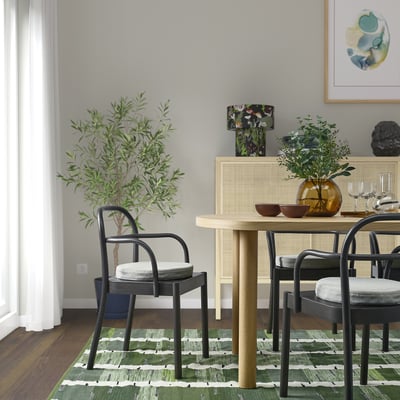 Modern dining area with black chairs, light wood table, green patterned rug, and decorative plants. Painting on wall, curtains on window.