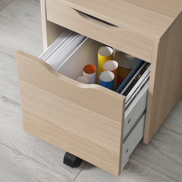 A light wooden desk with an open drawer holding coloured cylindrical containers, likely ALEX series, offering organised storage.
