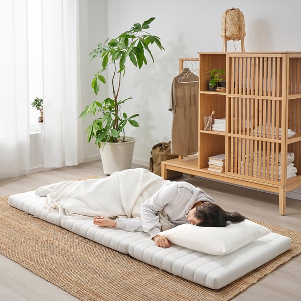 Living room: person sleeps on an ÅNNFJÄLLET white floor-mattress, with bookshelf, plant, lamp.