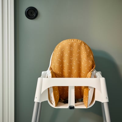A white high chair with a yellow and white spotted cushion provides stable support for a baby, attached to a green wall.