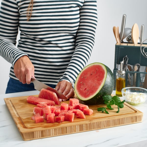 Person slicing watermelon on APTITLIG bamboo chopping board with grooves for juice collection, stable and ergonomic.
