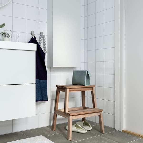 Modern bathroom with white tiles, white cabinet, dark blue towel, wooden step stool, blue bath bomb, slippers.