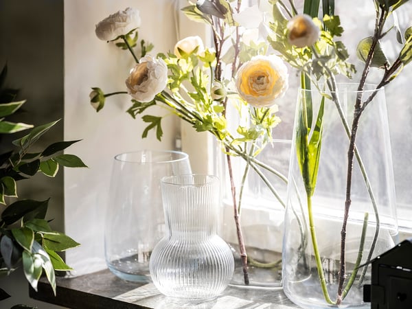 Clear glass vases with textured patterns hold light-colored flowers and green leaves, arranged on a windowsill.