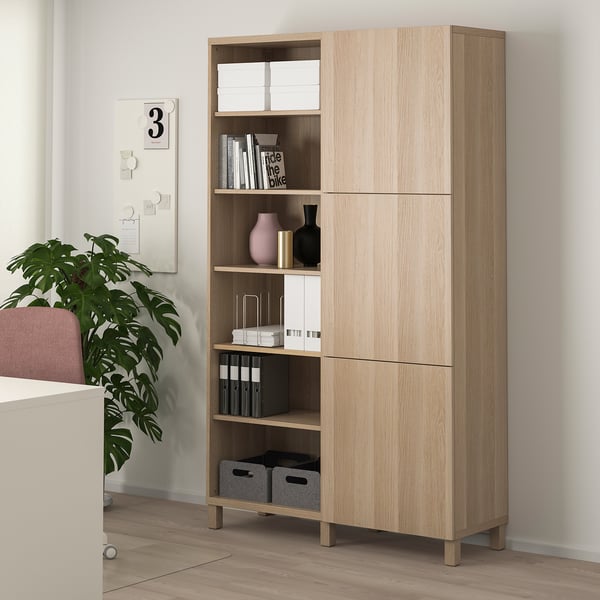 Light wood BESTÅ cabinet with open and closed storage, displaying books, vases, and boxes. It stands taller than nearby house plant and desk.