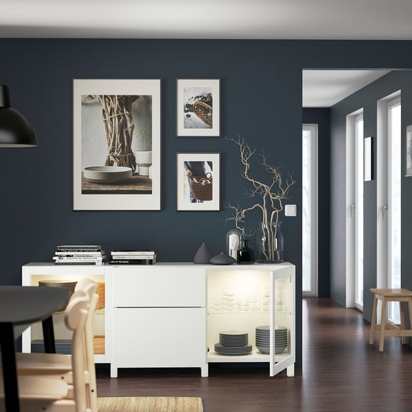 Modern dining room with dark walls, white sideboard featuring adjustable shelves and glassware, with framed artwork and stylish decor.