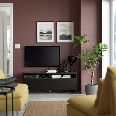 Living room with tv and yellow couch. BESTÅ tv stand in black, mounted on purple wall. Two framed nature pictures above, tall plant to right.