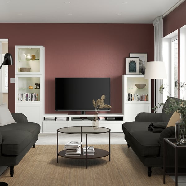 Modern living room with burgundy walls, grey couches, glass coffee table, BESTÅ tv unit, flat-screen tv, floor lamps, and potted plant by window.