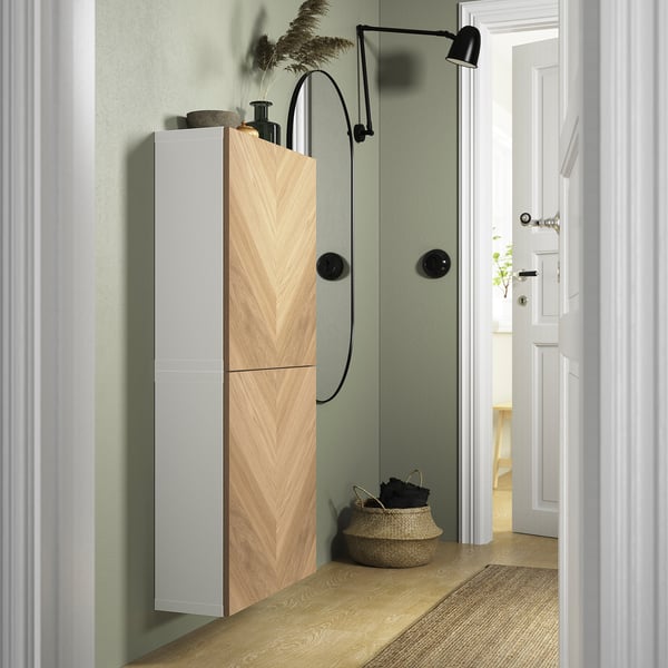 Modern bathroom with tall, sleek light wood BESTÅ cabinet, black accent wall lamp, and wicker basket beside a white door.