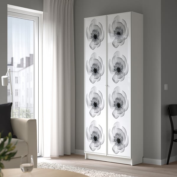 A tall, white BILLY bookcase by a window with flower designs, next to a curtain and chair.