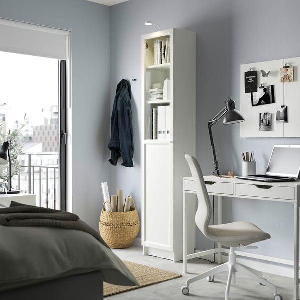Modern home office with BILLY bookcase, desk, and grey chair. Bookcase has glass doors.