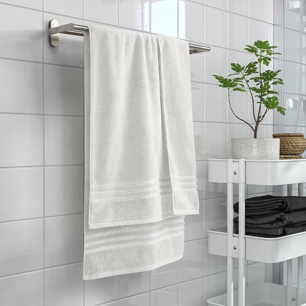 White BROKGLIM organic cotton towel on wall, larger than folded towels on shelf with plant.