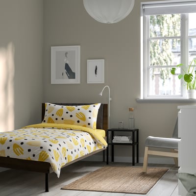 Bright bedroom with yellow-patterned bedding, grey walls, large window, and wall art. The bed is the centrepiece, topped with a colourful bedspread.