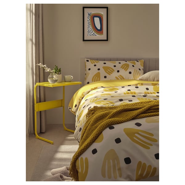 Bright bedroom with yellow patterned bedding, yellow table, and plant.