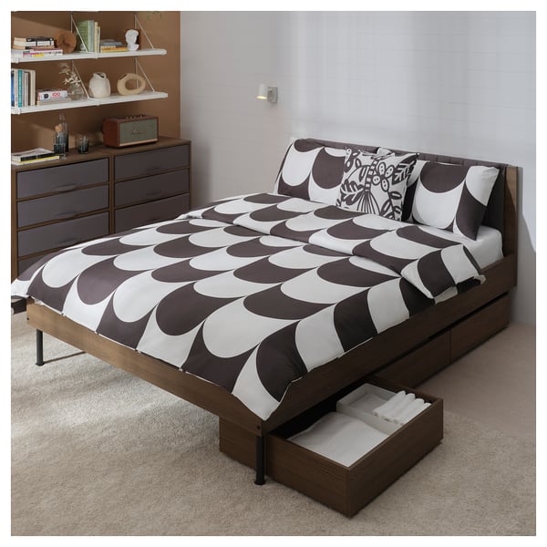 A BRUKSVARA bed in brown with a patterned comforter and cushions, featuring a soft headboard and storage boxes underneath.
