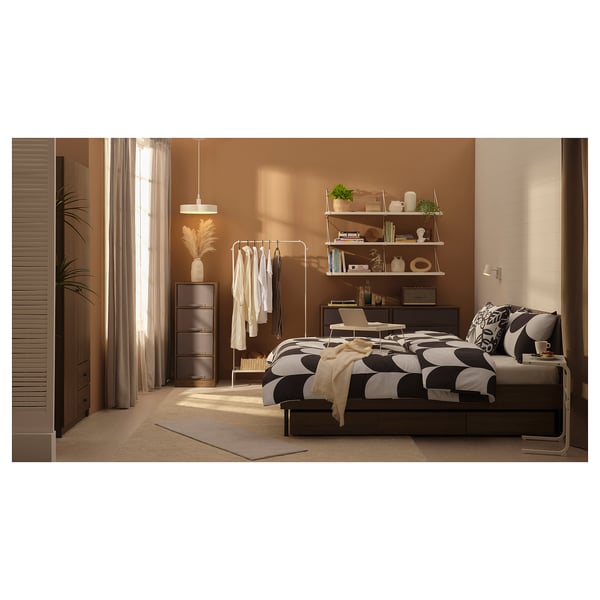 Bedroom with brown BRUKSVARA bed, patterned duvet. Wooden drawers and shelves, beige curtains.