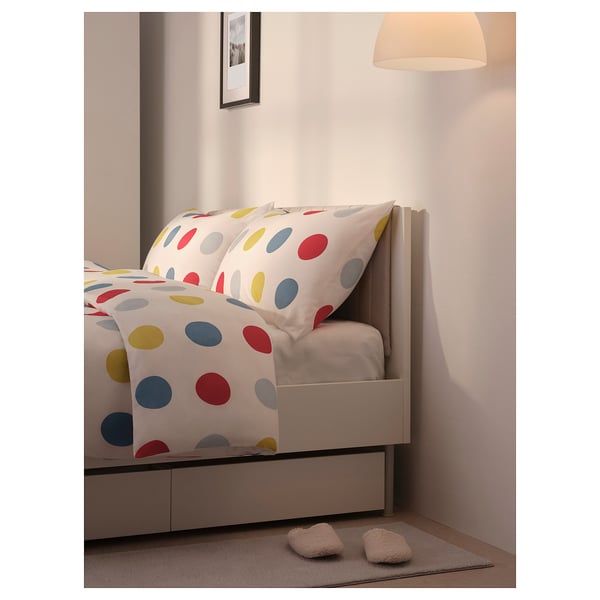 Bright polka dot bedding on white bed frame with cushioned headboard and wall lamp.