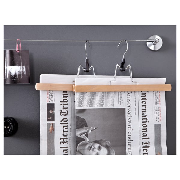 Clothes hanger with wooden bar and metal hooks holding newspaper against grey wall.