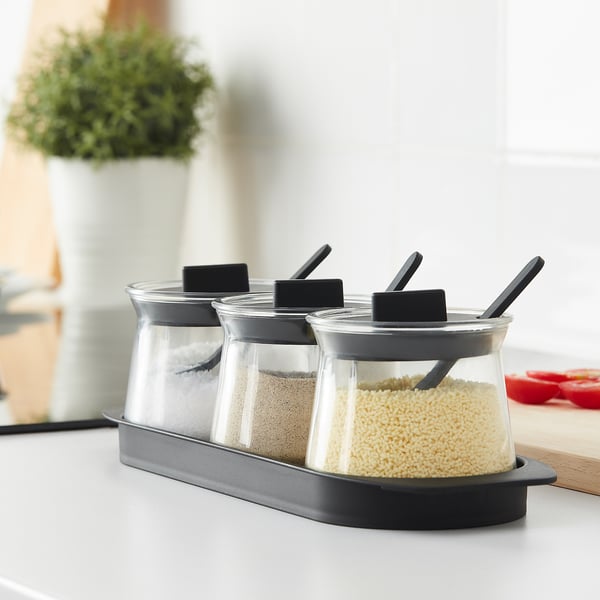 Three clear glass spice jars with black lids and spoons on a black tray, each containing different spices, for easy storage and access.