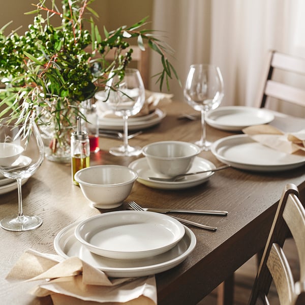 DAGGASTER stoneware in light beige, irregular speckles, set on table with wine glasses, napkins, and greenery.