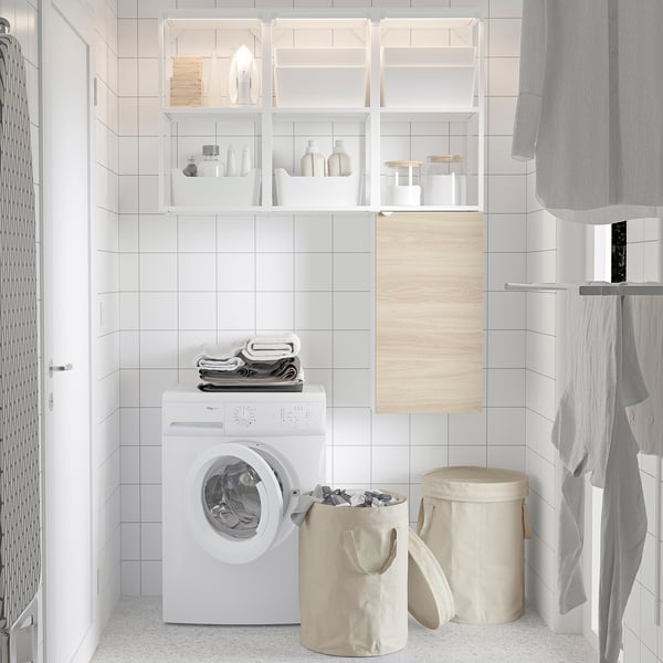 Laundry room with washing machine, clothes, and items on shelves. Beige DIMPA bag with lid and divider near cabinet and towels.