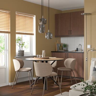 Cosy dining area with DOCKSTA round table, four chairs, and pendant lights. Kitchenette and window in background.