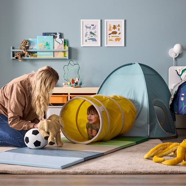 A kid plays inside a yellow tunnel, mum bends to greet, football ball and teddy bear beside, blue tent nearby, books and toys on shelf.