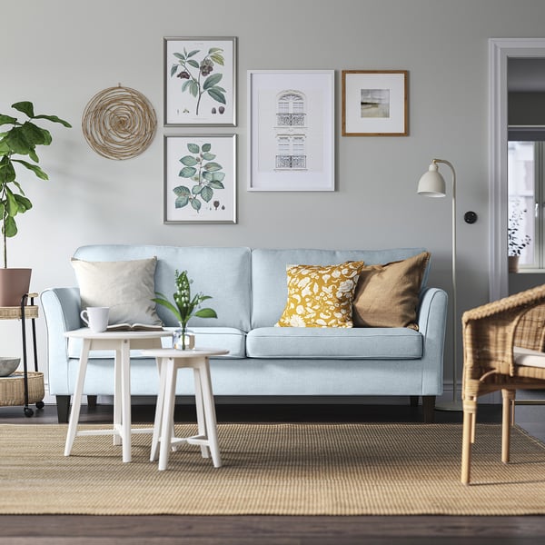 Cosy living room with light blue EKHOLMA sofa, colourful pillows, wicker chair & plant. Gallery wall, floor lamp. Modern & inviting.