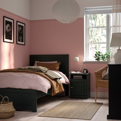 Modern bedroom with dark wood bed, pink walls, flowers, wicker chair, & storage.
