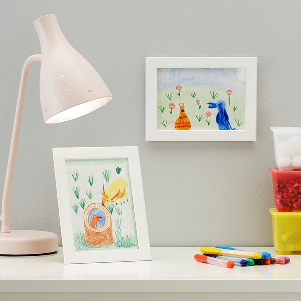 Desk with lamp, framed drawings, markers, and containers. Lamp illuminates workspace.