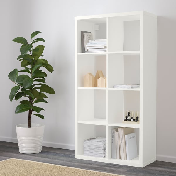 A white shelving unit, standing vertically, holding books and decorative items on its shelves. A potted plant is placed nearby on the floor.