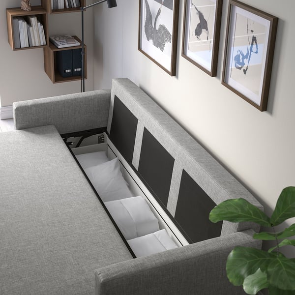 Modern sofa with open storage, showcasing bedding inside. Art frames on wall, plant at right.
