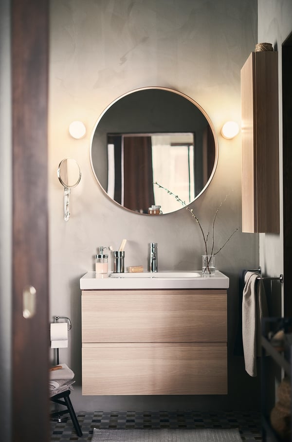 Modern bathroom with round mirror, light wood vanity, white sink, metallic fixtures, and glass vase with branches.