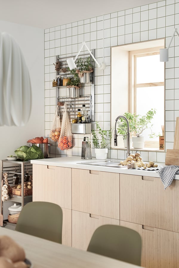 Modern kitchen with light wood cabinets, white tiles, hanging plants, and fresh produce on counter.