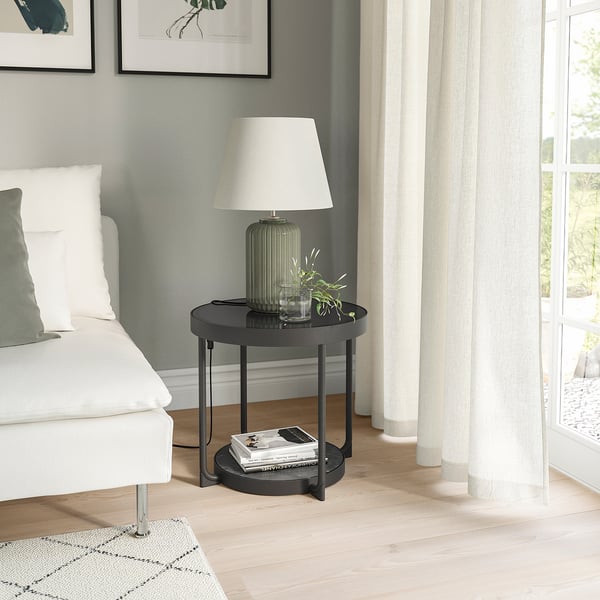 Modern black side table with smoked glass top, marble shelf, lamp, books, and plant beside bed. Contemporary design.